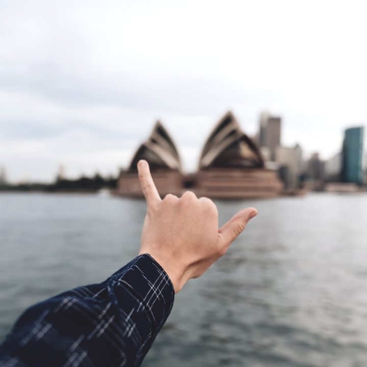 Serviços de Acomodação para Intercâmbio. Foto com mão de pessoa fazendo sinal em frente ao Opera House, em Sydney, na Austrália.